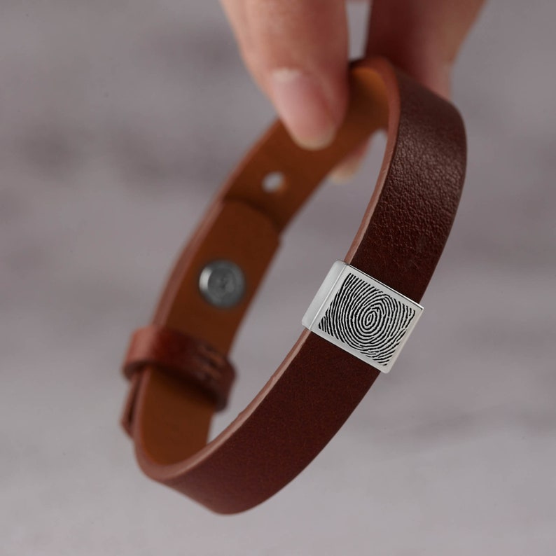 May include: A brown leather bracelet with a silver clasp displaying a fingerprint. The bracelet, with a magnetic clasp, is held up against a blurred background.