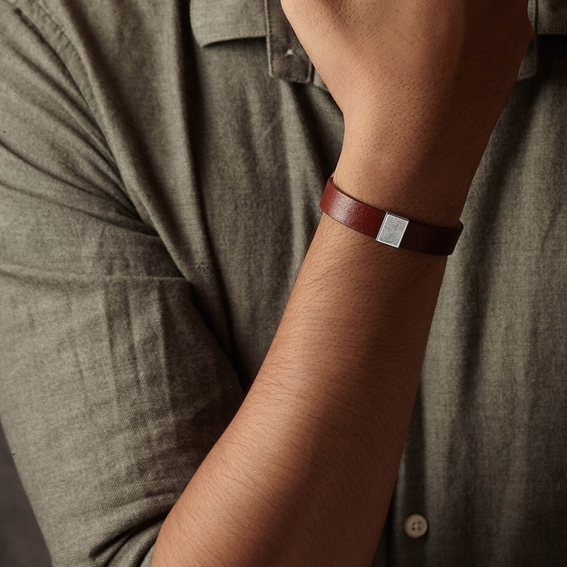 May include: A brown leather bracelet with a silver rectangular clasp. The bracelet is worn on a person's wrist, against the backdrop of a green shirt.