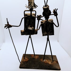 May include: A metal sculpture of two figures, one holding a walking stick, standing on a black metal base. The figures are made of wire and metal scraps, and they are painted black.