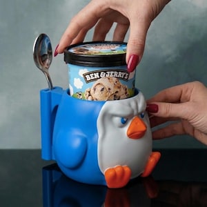 May include: A blue and white penguin-shaped ice cream pint holder with an orange beak and feet. The holder features a side slot for a silver metal spoon and contains a pint of Ben & Jerry's ice cream with a graphic of a scoop of cookie dough.