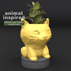 May include: A yellow geometric cat-shaped planter with a plant inside. The planter is on a gray base. The text "animal inspired" and "planter, pen holder, candy bowl" are in the image.