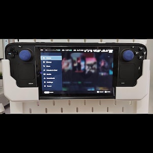 May include: A black handheld gaming console with blue thumbsticks is mounted in a white holder. The screen displays a menu with options like "Home," "Library," and "Store." The console is attached to a white pegboard.