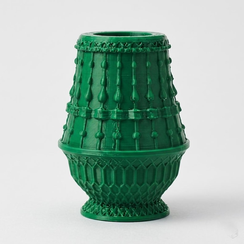 May include: A dark green vase with a tapered cylindrical top section featuring vertical raised lines and small bead accents, transitioning into a wider, bowl-shaped base with a detailed lattice or honeycomb pattern throughout the lower portion.