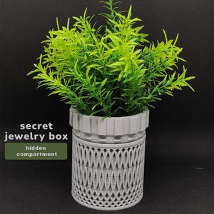 May include: A gray, decorative jewelry box with a hidden compartment, designed to look like a potted plant. The box has a textured, woven pattern on the lower section and a ridged top. The plant has bright green leaves. Text on the image reads "secret jewelry box hidden compartment."