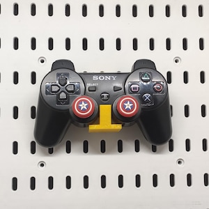 May include: Black Sony PlayStation controller with red, white, and blue Captain America button covers. The controller is mounted on a yellow stand against a white pegboard background.