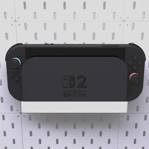 Puede incluir: Una consola y una base Nintendo Switch negras están montadas en una pared perforada blanca. La consola tiene mandos azules y rojos. La base muestra el logotipo de Nintendo Switch. La pared perforada tiene agujeros espaciados.