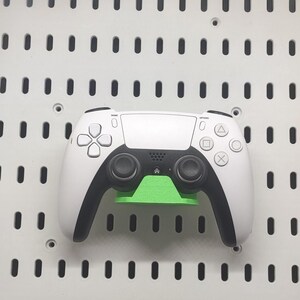 May include: A white and black video game controller with a green stand, mounted on a white pegboard. The controller has black handles and buttons with a directional pad and action buttons.