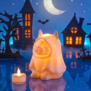 Pode incluir: Uma luz noturna em forma de gato com tema de Halloween que emite um brilho laranja quente. O gato está usando uma fantasia com capuz e tem um rosto sorridente. Uma pequena vela acesa está colocada em frente ao gato. O fundo mostra uma cena assustadora com casas, uma lua e morcegos.