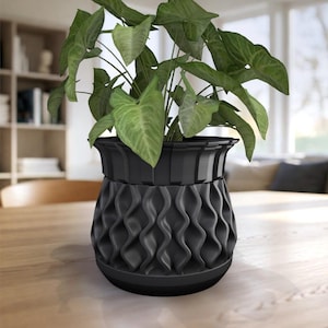 Puede incluir: Una maceta de color negro mate con un patrón texturizado de costillas verticales onduladas en el exterior y un borde liso y acampanado en la parte superior. La maceta contiene una planta verde con follaje en forma de corazón.