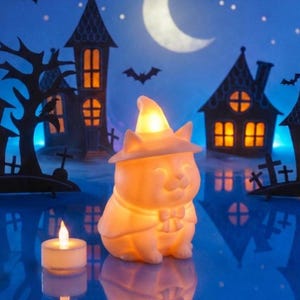 Peut inclure: Une figurine de chat d'Halloween lumineuse portant un chapeau de sorcière et une cape. Le chat est éclairé d'une lumière chaude, avec une petite bougie à proximité. Le fond présente des maisons effrayantes, des chauves-souris et un croissant de lune.