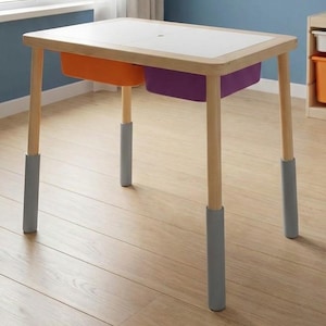 May include: A small wooden table with a white top and two storage bins, one orange and one purple. The table has light-colored wooden legs with gray protective sleeves. The table is designed for children.