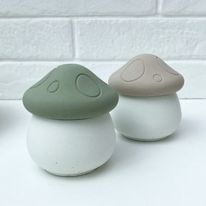 May include: Two small ceramic mushroom-shaped containers with white bases and green and brown tops. The containers are sitting on a white surface.