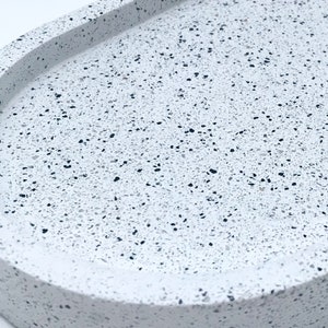 May include: A white oval tray with a speckled terrazzo pattern. The tray has a raised rim and a smooth, slightly rounded surface.
