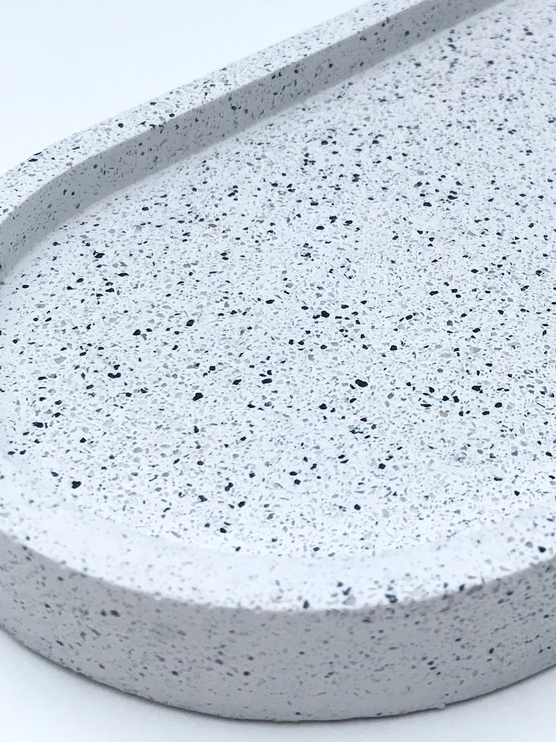 May include: A white oval tray with a speckled terrazzo pattern. The tray has a raised rim and a smooth, slightly rounded surface.