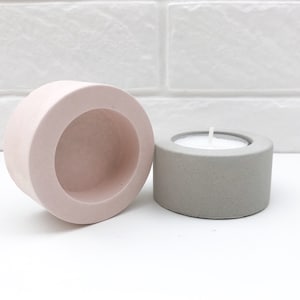 May include: Two concrete candle holders, one in a light pink colour and the other in a light grey colour. Both holders have a round opening in the centre for a tea light candle.
