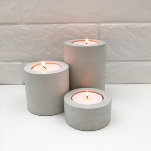 May include: Three grey concrete candle holders with lit tea lights. The candle holders are different sizes and are arranged in a row on a white surface.