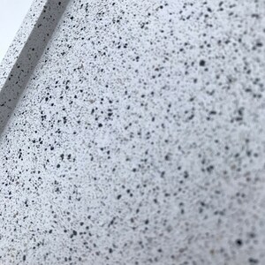 May include: Close-up of a white surface with small black flecks. The surface has a rough texture.