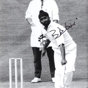 Op de afbeelding: Zwart-witfoto van een cricketwedstrijd. Een bowler in een wit uniform is in actie, terwijl hij de bal gooit. Een scheidsrechter staat achter hem. Cricket stumps zijn zichtbaar op de voorgrond.