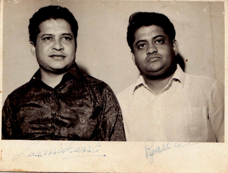 May include: Black and white photo of two men standing side by side. The man on the left is wearing a patterned shirt and the man on the right is wearing a white shirt. Both men are smiling and have their signatures below their images.