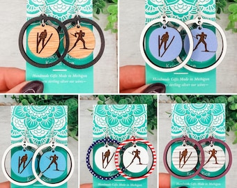 May include: Five pairs of dangle earrings featuring circular frames and inner discs with laser-etched skier silhouettes. The designs include natural wood, light blue, white, and striped patterns, all attached to sterling silver ear wires on display cards.