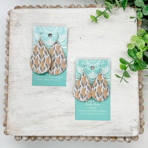 May include: Teardrop-shaped earrings with a corn-like pattern in yellow, gray, and white, displayed on a white wooden surface. The earrings are on turquoise cards with text. The earrings are silver-toned.