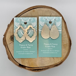 May include: Two pairs of gold-toned earrings on a teal and white card with the text "Fleece & Fancy Kristin Rice Handmade Gifts Made in Michigan kristin@fleeceandfancy.com".