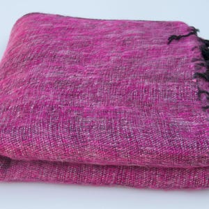 May include: A folded rectangular throw blanket featuring a vibrant magenta and black woven texture. The fabric displays a dense, heathered appearance with a mix of dark and bright fibres throughout. One edge is finished with short black fringe tassels.
