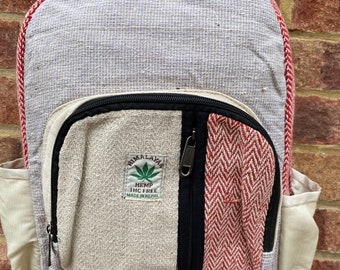 May include: A hemp backpack with a multi-coloured design. The main body is light grey with red trim. The front pocket features a cream-coloured section, a red and white chevron pattern, and a black zip. The text "Himalayan Hemp THC Free Made in Nepal" is visible.