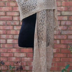 May include: A light brown, open-knit shawl with a geometric design, displayed on a black mannequin. The shawl features a fringed edge and is set against a red brick wall. The knitted pattern is visible throughout the fabric.