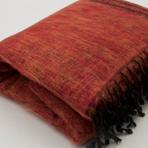 Könnte beinhalten: Eine rote und orangefarbene Wolldecke mit schwarzen Fransen. Die Decke ist in der Hälfte gefaltet und die Fransen sind auf beiden Seiten sichtbar.
