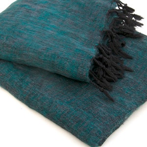 May include: Folded teal and grey woven blanket with black fringe. The blanket has a textured appearance and is made of a soft material. The fringe is thick and adds a decorative element to the blanket.