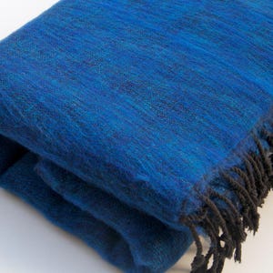 Puede incluir: Un chal de lana azul con flecos negros. El chal está doblado y tiene una textura suave y peluda.