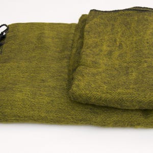 Pode incluir: Cobertor verde-oliva dobrado com franjas pretas. O cobertor tem uma trama texturizada e uma borda escura. As franjas s&atilde;o amarradas e adicionam um elemento decorativo. O cobertor &eacute; dobrado cuidadosamente, mostrando sua textura e cor.