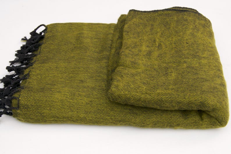 Pode incluir: Cobertor verde-oliva dobrado com franjas pretas. O cobertor tem uma trama texturizada e uma borda escura. As franjas s&atilde;o amarradas e adicionam um elemento decorativo. O cobertor &eacute; dobrado cuidadosamente, mostrando sua textura e cor.