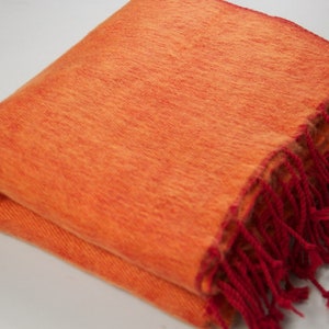 Puede incluir: Una manta suave de color naranja con flecos rojos. La manta está doblada por la mitad y tiene una superficie texturizada.
