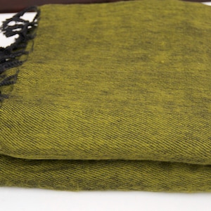 Pode incluir: Cobertor verde-oliva dobrado com franjas pretas. O cobertor &eacute; feito de um tecido texturizado e &eacute; dobrado cuidadosamente. A franja est&aacute; ao longo de uma borda do cobertor. O cobertor est&aacute; em uma superf&iacute;cie branca.