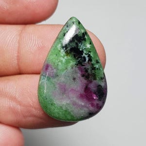 Può includere: Una gemma a forma di goccia lucidata. La pietra presenta un mix di colori verde, nero e magenta. Il verde è il colore dominante, con inclusioni nere e magenta. La gemma è tenuta in una mano.