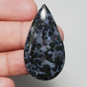 May include: A teardrop-shaped gemstone with a polished surface. The stone features a mottled pattern of dark blue, black, and light blue hues. The gemstone is held in a hand, showcasing its size and shape.