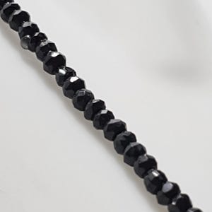 Peut inclure: Gros plan sur un collier de perles noires. Les perles sont facettées, réfléchissant la lumière, et enfilées pour former une chaîne délicate. Le collier est présenté sur fond blanc, soulignant sa couleur sombre et son design complexe.