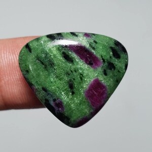 Könnte beinhalten: Ein polierter Edelstein mit dreieckiger Form. Der Stein hat eine leuchtend grüne Grundfarbe, gesprenkelt mit schwarzen und tiefroten Einschlüssen. Der Edelstein wird von einem Finger gehalten, der seine glatte, reflektierende Oberfläche zeigt.