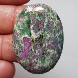 May include: An oval-shaped polished gemstone featuring a mottled pattern of vibrant magenta-pink, deep forest green, and creamy white mineral inclusions. The surface is smooth and reflective, showcasing a natural, variegated texture throughout the stone.