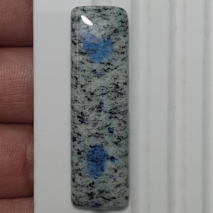May include: A rectangular K2 stone cabochon with a polished, rounded top. The stone features a speckled pattern of black and gray, with two prominent blue patches. The cabochon is approximately 1 inch long.