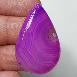 Peut inclure: Une pierre précieuse polie en forme de goutte d'eau, dans des tons de violet et de magenta. La pierre présente des motifs de bandes concentriques et une zone translucide près du sommet. Idéal pour la joaillerie.