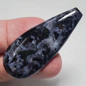 May include: A polished, teardrop-shaped gemstone with a dark blue and black mottled pattern. The stone has a glossy finish and is held in a hand, showcasing its natural beauty. This unique stone could be used for jewelry making.