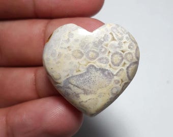 May include: A heart-shaped stone with a unique pattern of beige, white, and lavender hues. The stone's surface features a textured design with circular and irregular shapes. The heart is held in a hand, showcasing its polished finish and natural beauty.