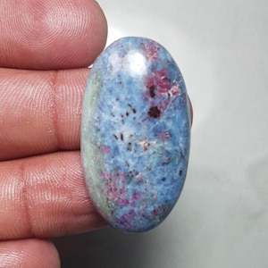 Könnte beinhalten: Ovaler Edelstein mit blauer Grundfarbe, gesprenkelt mit Weiß und Flecken in Rot und Dunkelbraun. Der polierte Stein wird in einer Hand gehalten, wodurch seine glatte Oberfläche und natürlichen Muster hervorgehoben werden. Ideal für die Schmuckherstellung.
