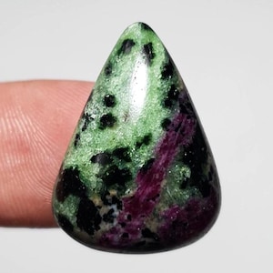 May include: A polished, teardrop-shaped gemstone with a mix of colors. The stone features a base of green speckled with black, and a streak of deep red-purple. The gem is held by a finger.