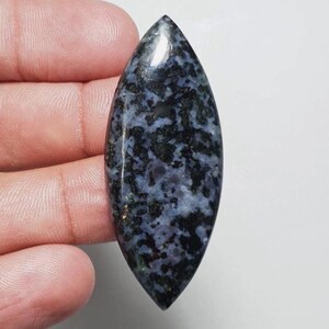 May include: A polished, elongated gemstone with a pointed tip. The stone features a mottled pattern of dark blue and black, with hints of lighter blue and green. The gemstone is held against a white background.
