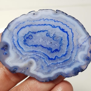 May include: A polished agate slice displaying concentric bands of white and blue hues. The natural stone features a central crystalline area and a smooth, rounded shape. The agate's unique patterns and colours make it a decorative item.