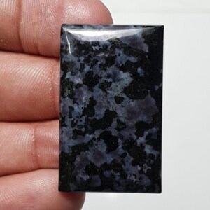 May include: A rectangular gemstone with a polished surface. The stone features a mottled pattern of black and blue-gray hues. The gemstone is held in a hand against a white background.